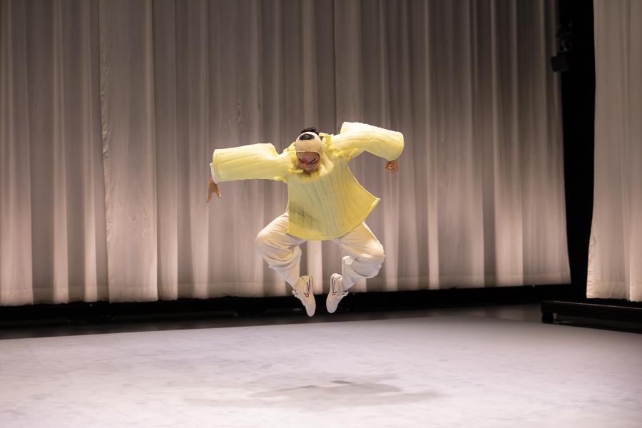 On a stage with a white curtain behind them, a performer, in a puffy yellow costume, leaps up with their toes pointed to each other.