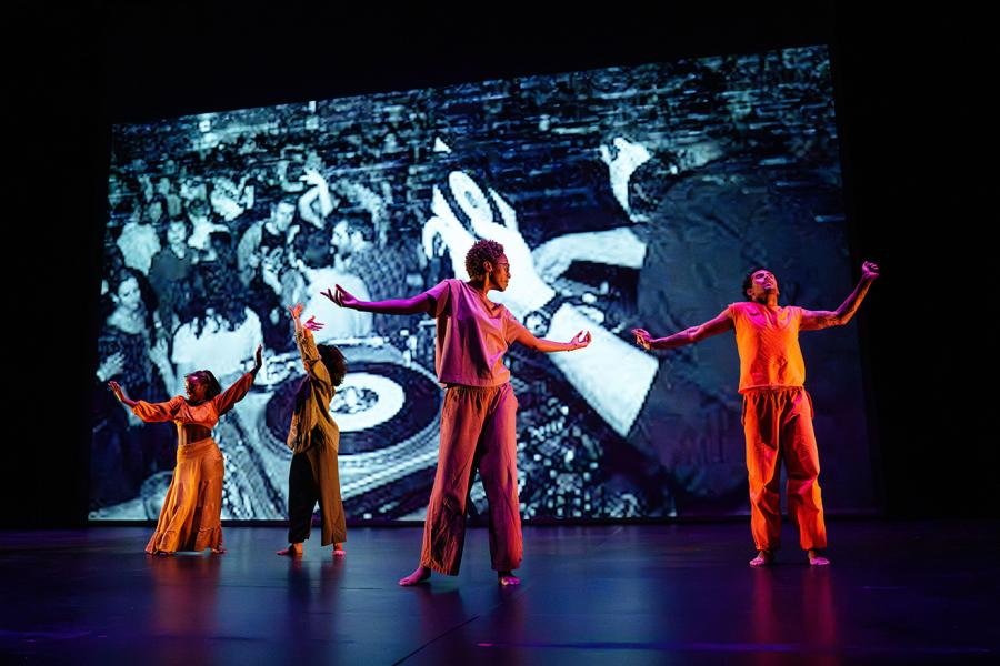 Four dancers stand in relaxed, open positions. Behind them is a projected archival photo of a DJ from the Shadow Lounge, a historic Pittsburgh club from the 2000s. One male-identifying dancer wears all orange; three female-identifying dancers wear yellow, green, and deep maroon. The group includes one Black man, two Black women, and one South Asian woman.