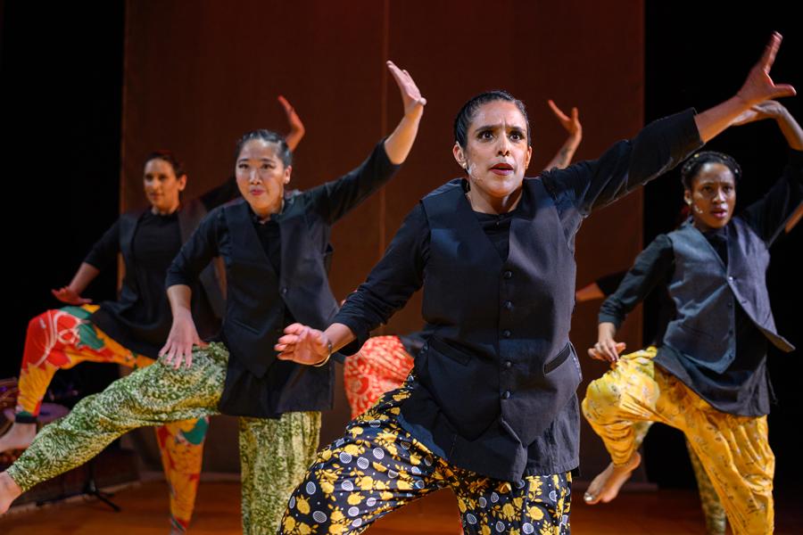 Four women of different races dance in black tops and printed bottoms, all different prints. They throw their left hands to the left and their right foot up.