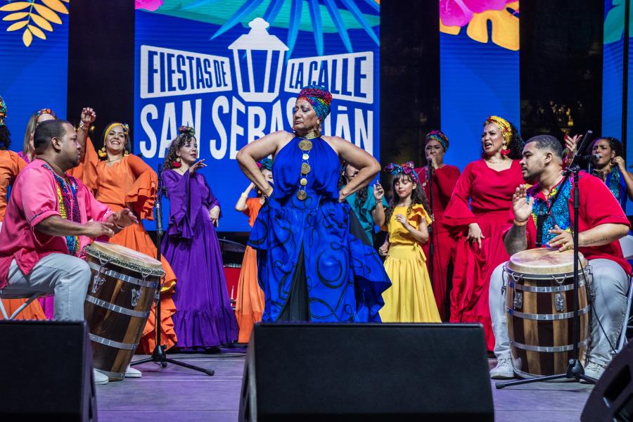 On a stage, a group of women perform at a fiesta. Two men play drums beside them.