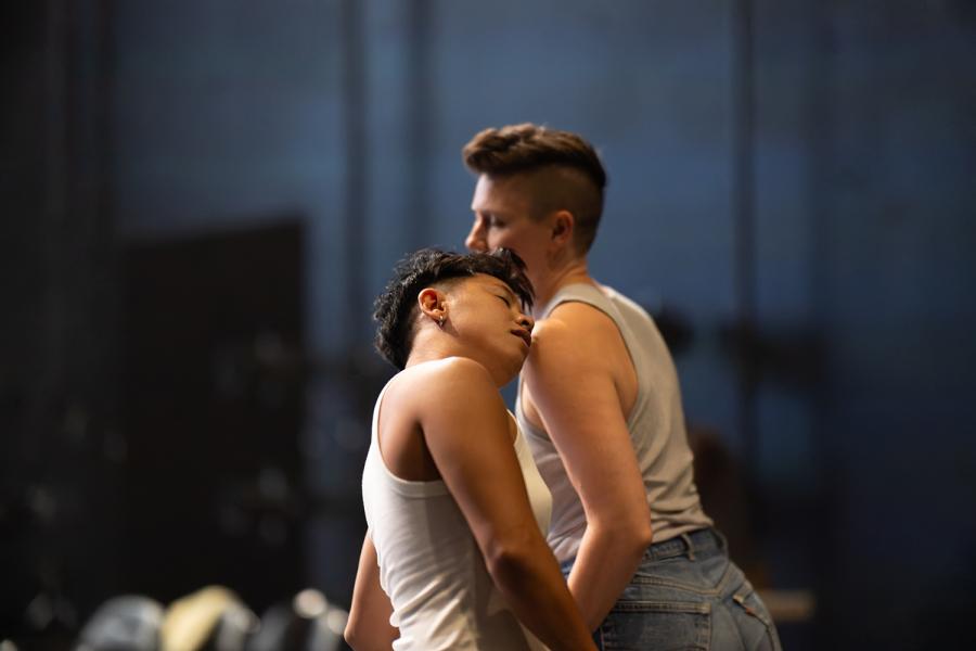 : Two dancers softly leaning on a diagonal at the center of the frame. Makisig Akin is in focus in front and Anya is blurry behind. Makisig’s eyes are softly closed. Both dancers are wearing light colored tank tops.