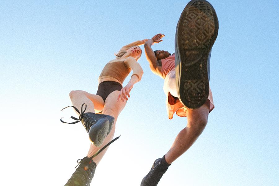 Two artists leap into the air and we see them from below their shoes in the expansive blue sky.