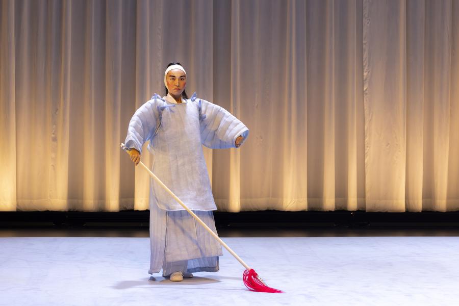 On a stage with a white curtain, a performer, in a clear mask and puffy blue costume, holds a white stick with red strings at the end in front of them.
