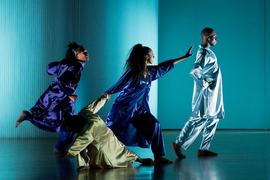 In a space with empty white walls and blue lights, four dancers in shiny gear walk towards the light. One falls but another holds their hand.