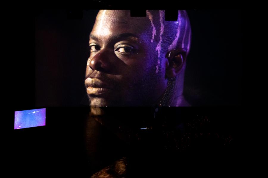 On a stage, a projection of jaamil's face with paint/makeup dripping down the back of their head. A tv with purple.