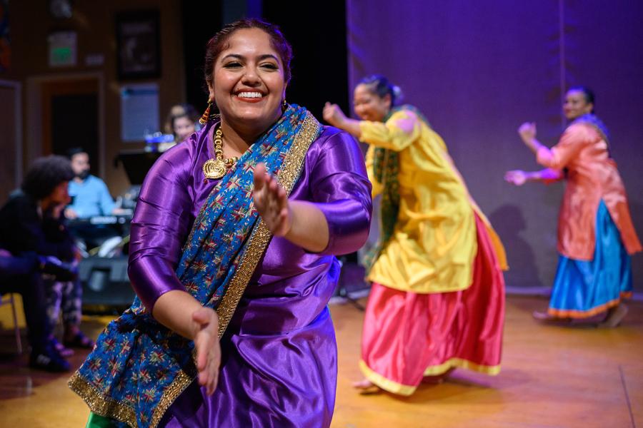 A brown woman smiles and claps while other performers dance behind her in traditional Indian garb.