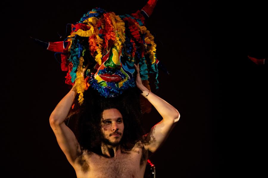Miguel holds a multi-colored, scrappy fabric mask over their head. They're shirtless and have brown hair to their shoulders.