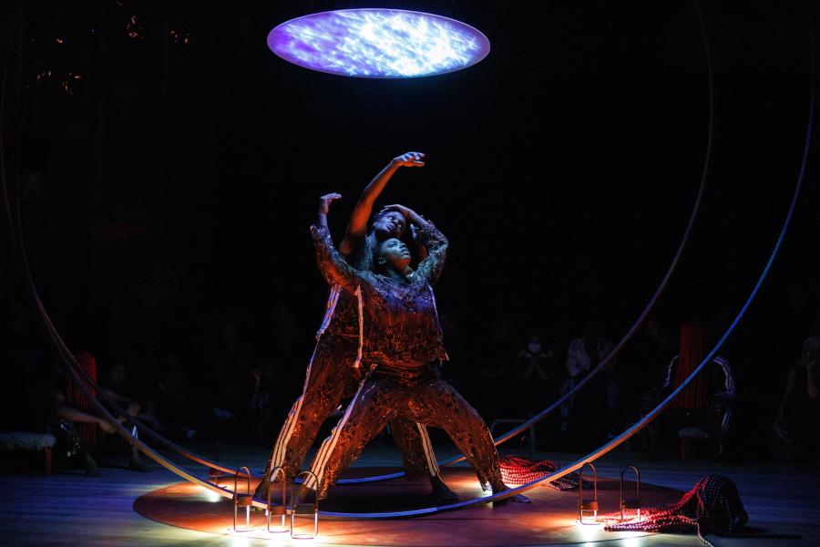 On a stage, between two large circular pole structures, and below a blue orb, two performers caress and look up at the orb.