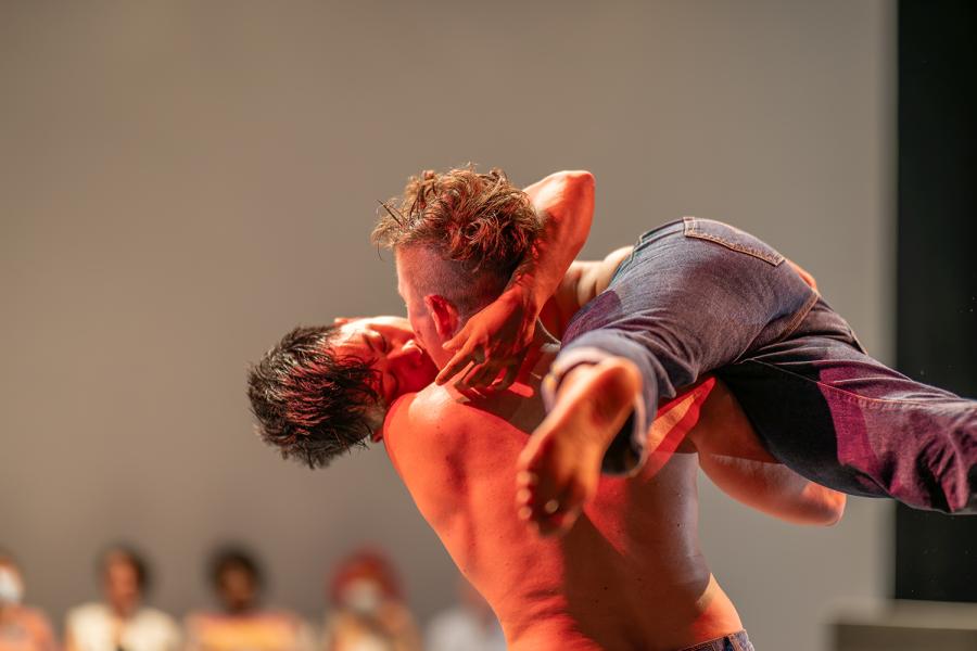 Two dancers kissing. Makisig Akin is lifted horizontally and balanced on Anya Cloud’s shoulder. Both dancers are shirtless and wearing jeans. Makisig is facing the camera with softly closed eyes. Anya is facing away from the camera.