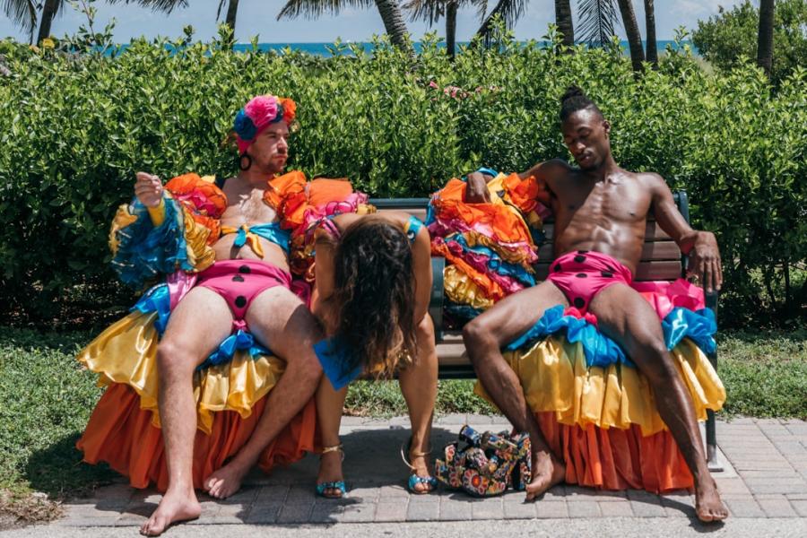 3 dancers in rumba costumes sit casually on a bench with the beach in the background. Their expressions range from exhaustion, disinterest and longing.