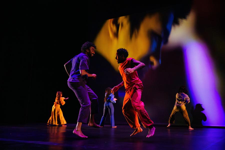 Five dancers are captured mid-motion. Two male-identifying dancers in the foreground wear purple and orange and move with energy. Behind them, three female-identifying dancers face away from the audience in a unified gesture. A club scene is projected in the background. The group includes two Black men, two Black women, and one South Asian woman. The dancers smile, radiating joy.