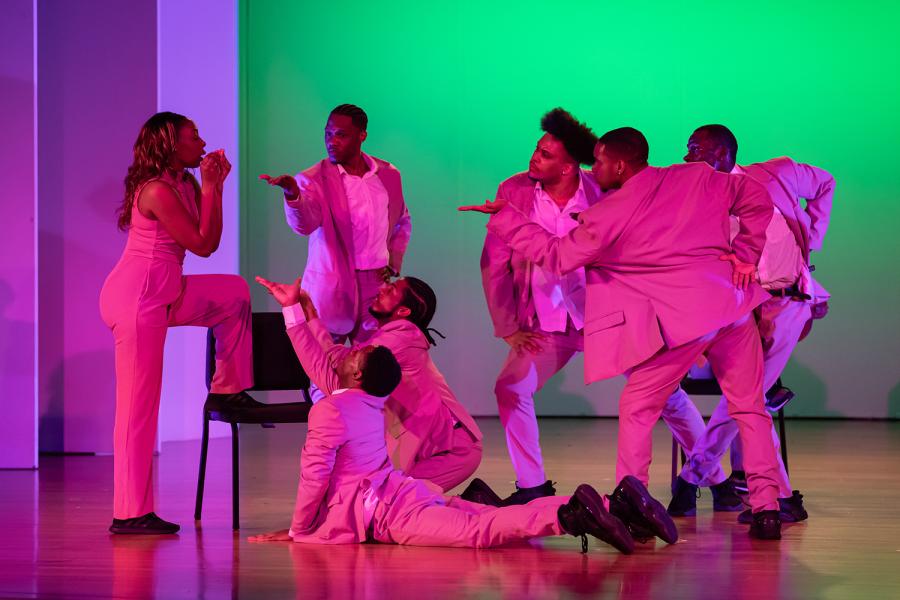 In pink glow, seven performers dance. One has her leg on a chair and faces the rest of the crew.