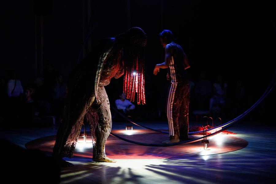 On a stage, between two large circular pole structures, two performs lean over, their breads catching lights from below them.