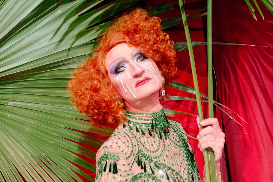 One performer in drag makeup and glitter tears holds palm fronds and stares into the camera with an expression of longing.