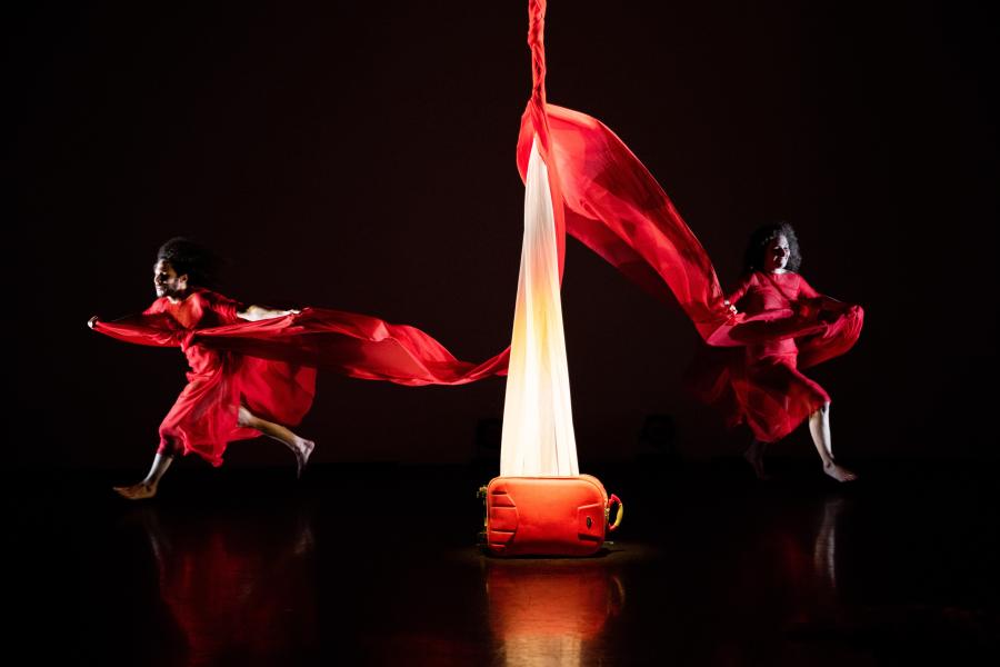 A backpack with light coming from it sits in the middle of a stage. Two performers, in red flowy gowns run in opposite directions from the middle.