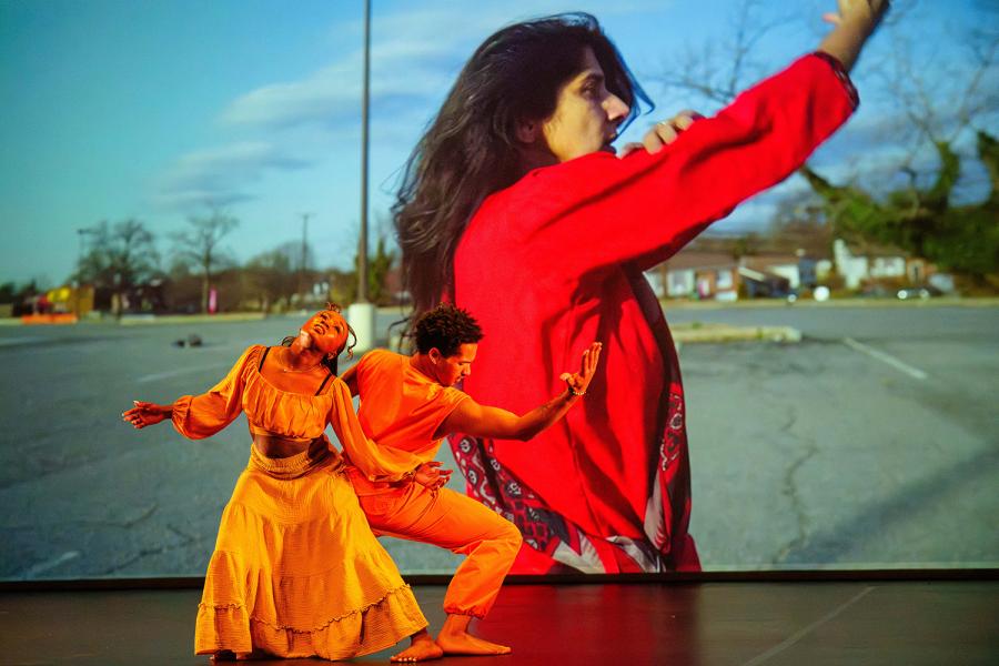 Two dancers perform a duet in physical contact, arms extended. The first is a Black female-identifying dancer in yellow; the second, a Black male-identifying dancer in orange. Behind them, a projected image shows a South Asian woman dancing alone in a parking lot, wearing a red traditional Indian salwar kameez.