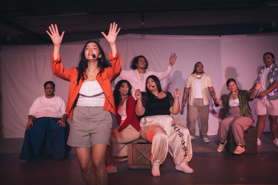 On a stage, a woman, in a red blazer, holds her hands up and speaks into a headset. Behind her, more performers hold their hands up, sit, or lean into each other.