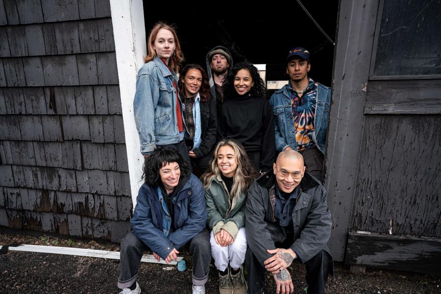 In a doorway, eight folks pose together in hoodies and denim coats.