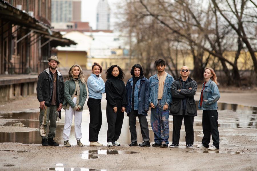 Outside, by some trees and a warehouse, eight folks stand and pose together.