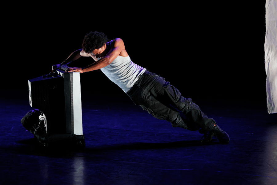 A performer in a white top and black jeans pushes a television on a dark stage.