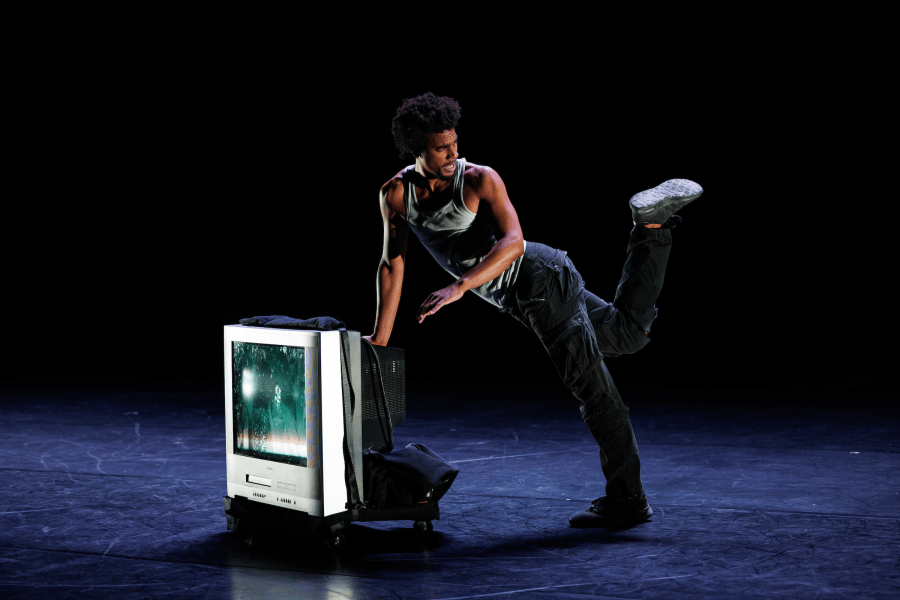 A performer kicks up a foot while leaning against a television box on a dark stage.