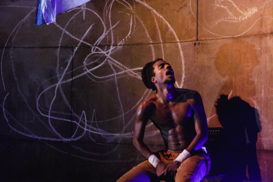 A performer in brown pants sits on a chair. Behind them, is a wall that has been chalked.