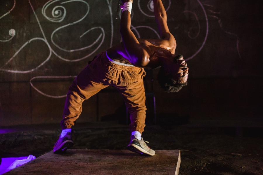A dancer wearing brown pants arches their spine backwards while throwing up their arms.