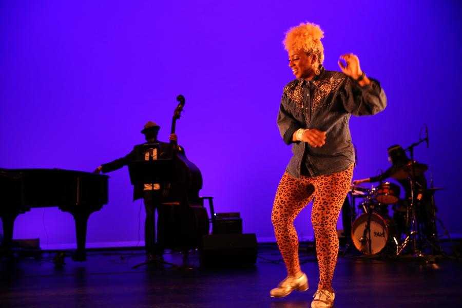 A Black woman, with a bleach blonde afro, dances in front of a band and a purple backdrop.