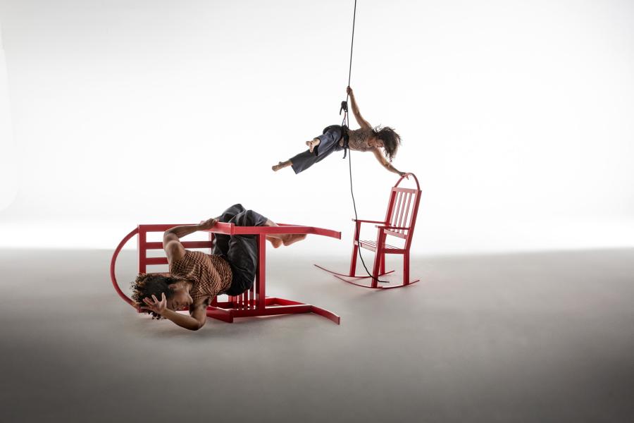 A person suspended in midair by a harness grabs a red rocking chair. A copy of the same person is shown wrapped around a flipped over red chair.