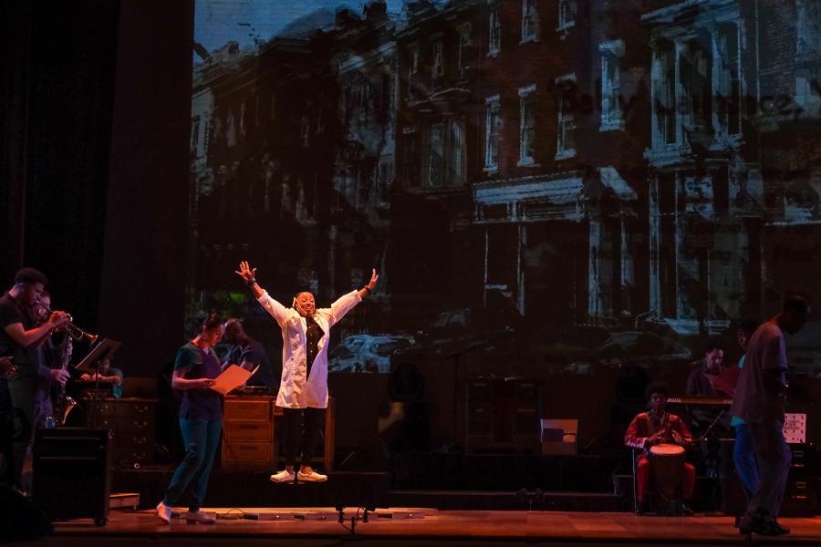 On a stage, a female tap dancer leaps straight up with her arms raised.  Behind her, a live jazz band and a projection of a city street.