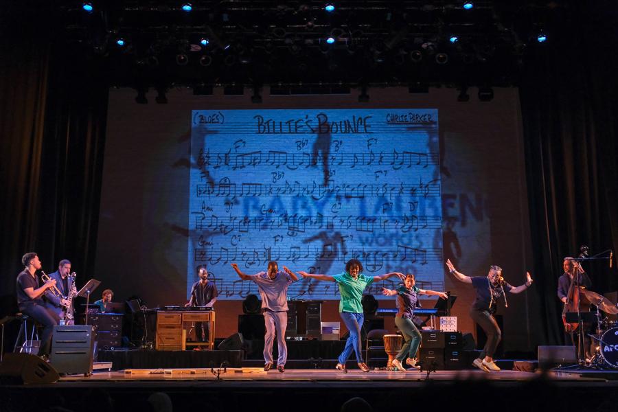 On a stage, a crew of tap dancers perform in front of a live band and a projection with musical notes and silhouettes of tap dancers.