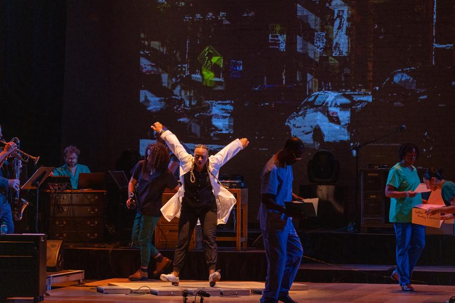 On a stage and in a spotlight, a female tap dancer hops onto her heels and raises her arms. A live band surrounds her.