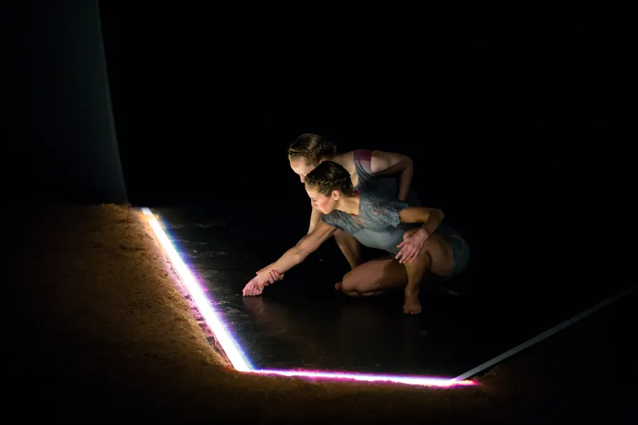 Two performers crouch by a bright tube of light and inspect it.