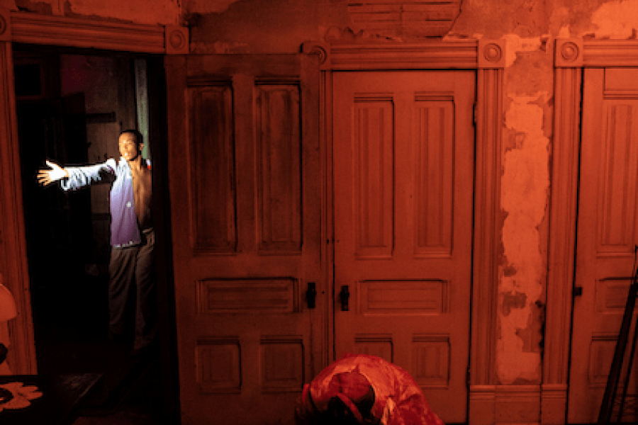 Inside a run down room, two people crouch on all fours. In the doorway someone stretches out an arm.