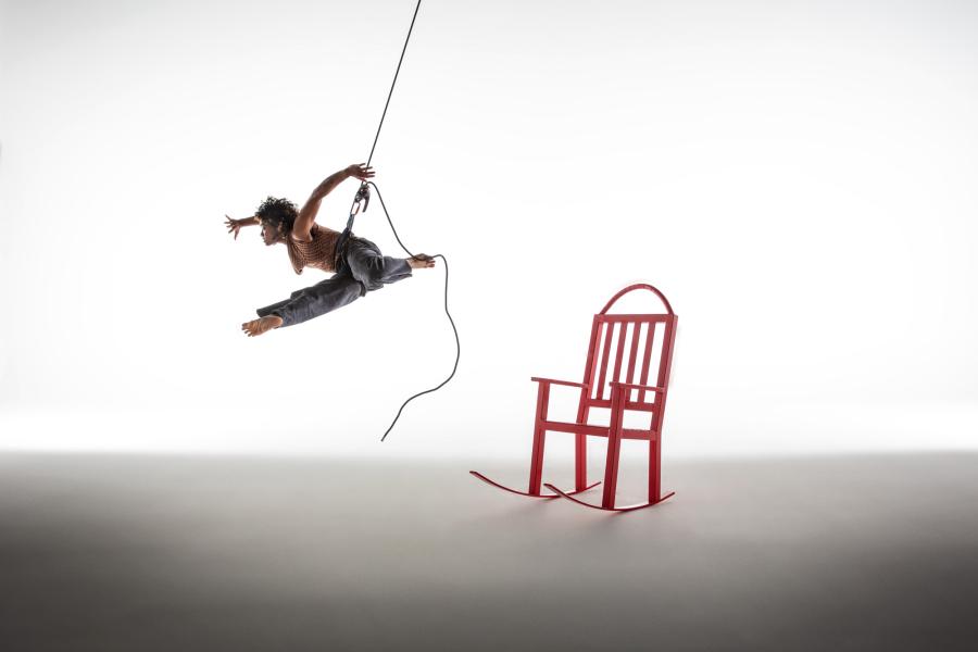A person suspended in midair by a harness in a white room with a red chair swings towards the right.