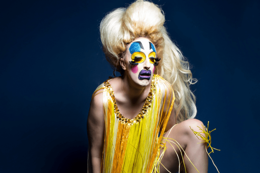 A drag queen with clown makeup poses in front of a blue background.