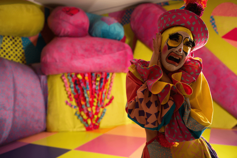 A drag queen dressed like a clown stands in front of a colorful background.