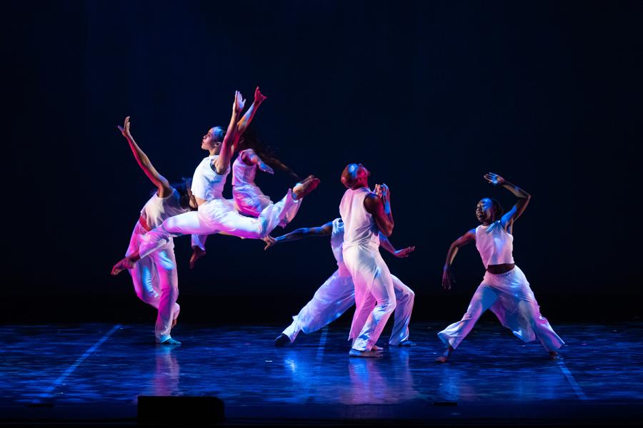 On a stage, six dancers, in all white costumes, perform. Two leap in the air, one in front of the other. 