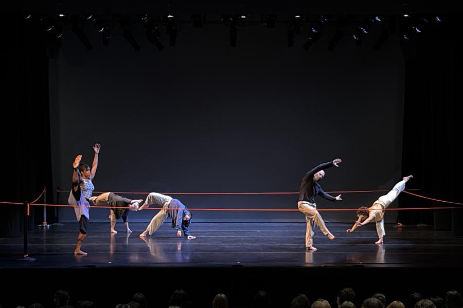 Five dancers strike poses on a stage that is illuminated by an overhead light.