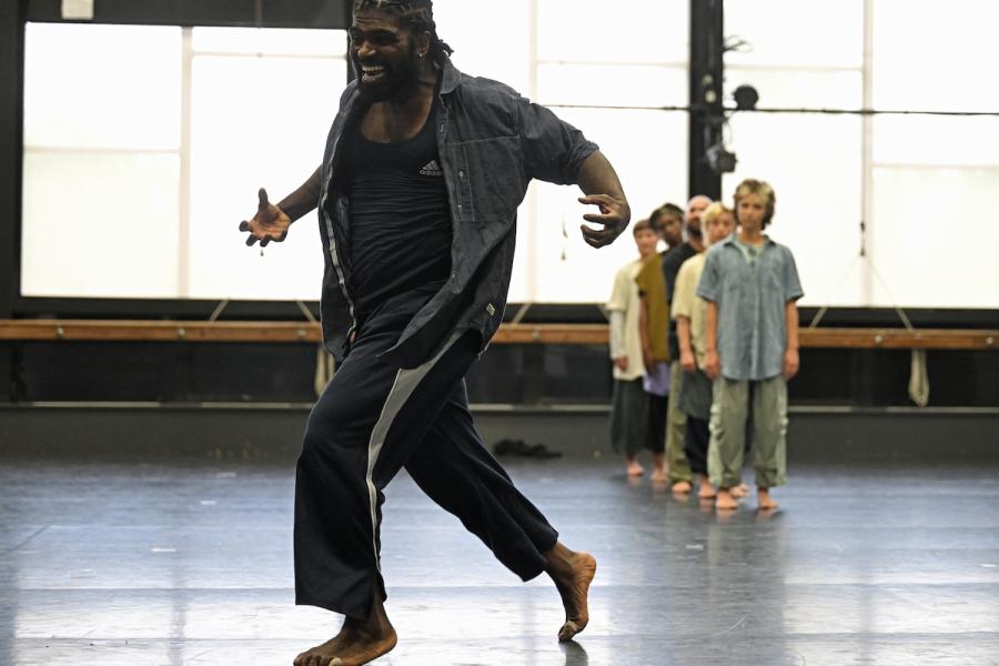 In the foreground a Black person strikes a passionate pose while in the background a group of five people standing in single file watch.