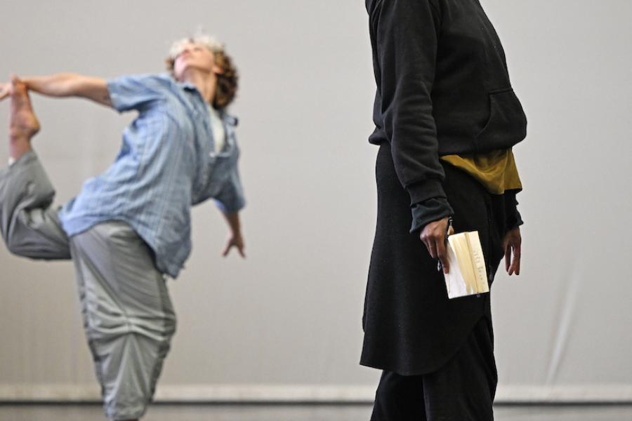 In the background a person dances on one foot, while the in the foreground a person stands sideways, looking off into the distance.