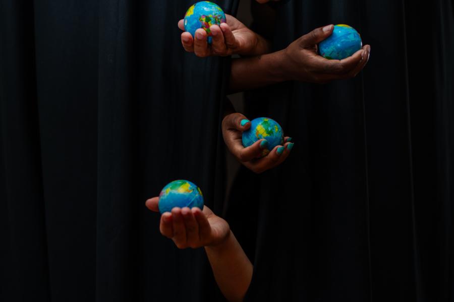Five hands poke out from a set of black curtains, each one holds a miniature globe.