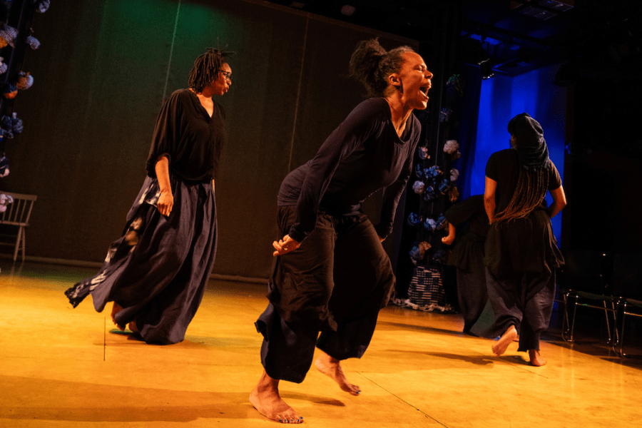 Four performers dressed in black move about a stage. One of them seems to scream passionately.