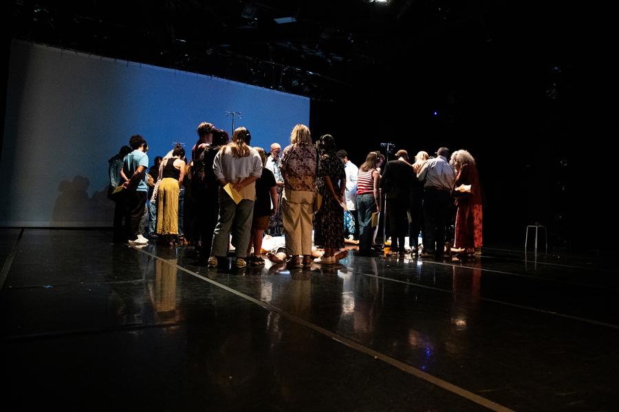 On a stage, a crowd, in plain clothes, surrounds dancers who lay flat on the stage.