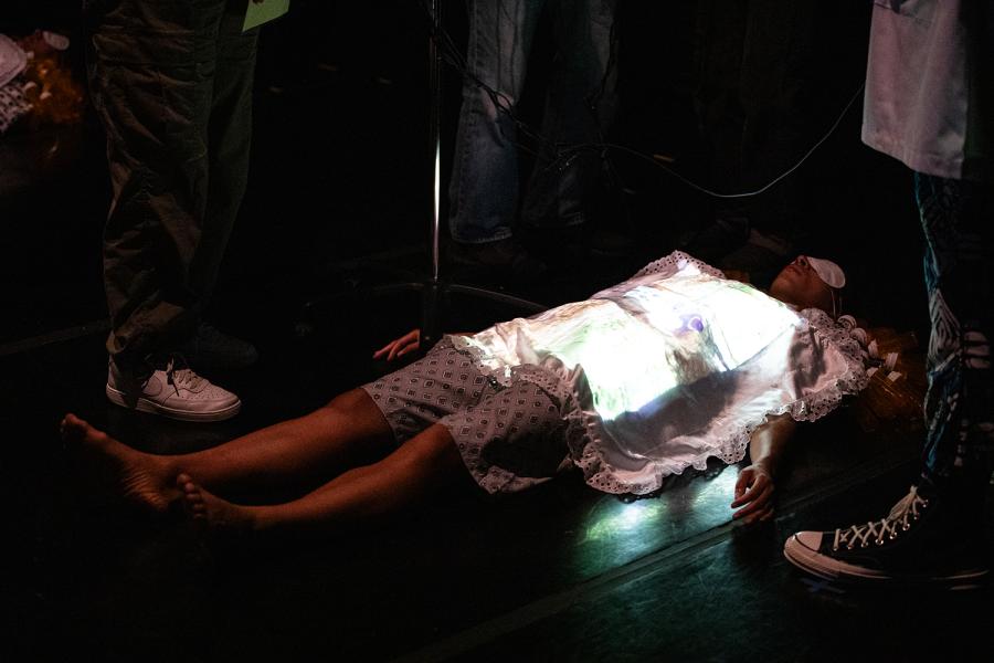 On a stage, a woman - in a spotlight - lays on the ground in a hospital gown and an eye mask. A small blanket covers her torso and she is surrounded by the audience who stands above her.