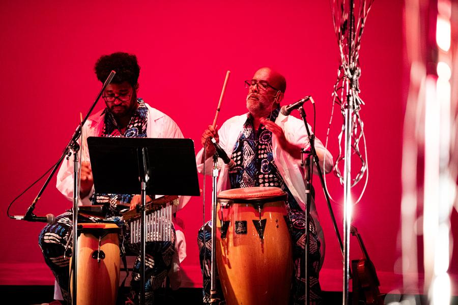 On a stage, with a red backdrop, men of color play the drums.