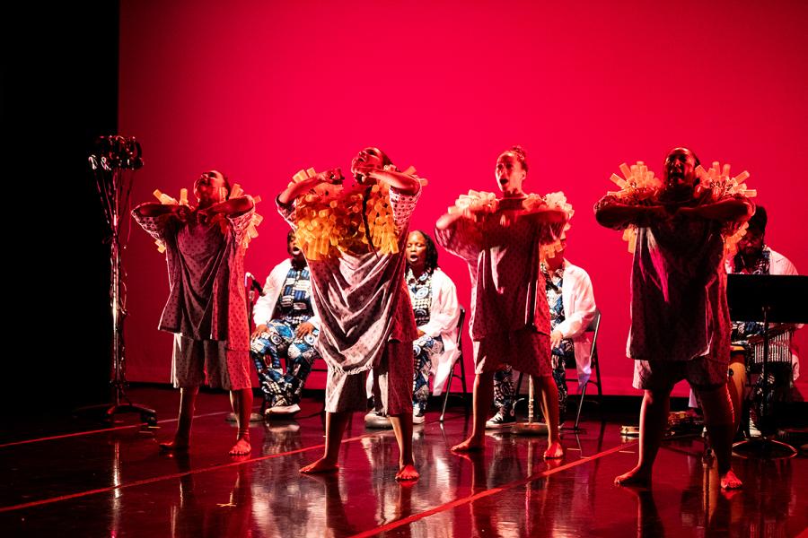 On a stage, with a red backdrop, four dancers in fuzzy costumes perform in front of a live band.