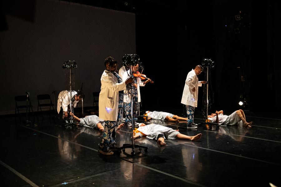 On a stage with a black backdrop, four folks lay on the ground in hospital gowns while four folks in doctor's coats inspect them from above.