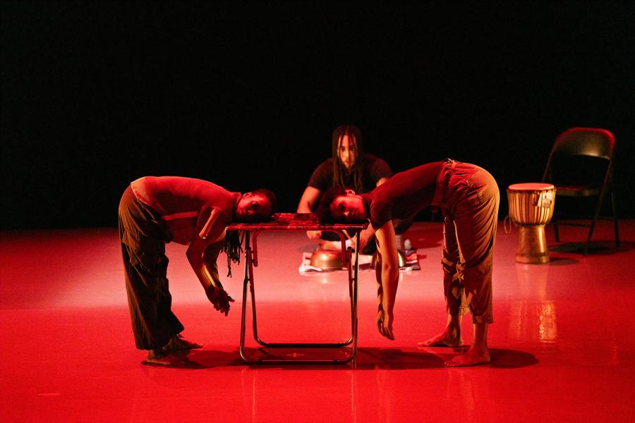 Two dancers lay their heads on a folding table, under red lights and on a stage. Another dancer is behind them as well as a bongo.