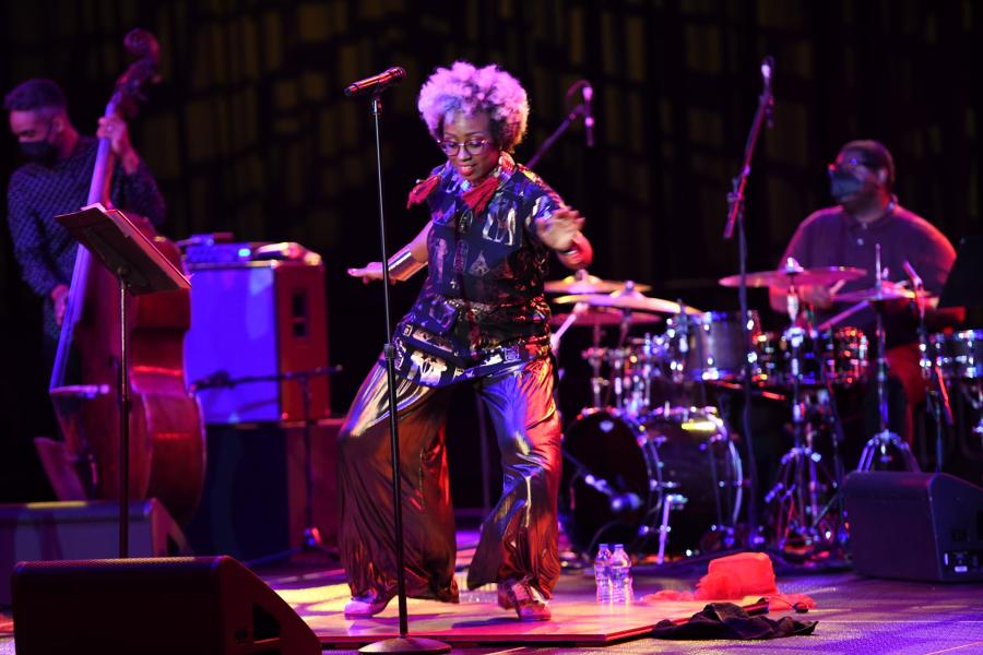 A Black woman, with a bleach blonde afro, dances in front of a band.